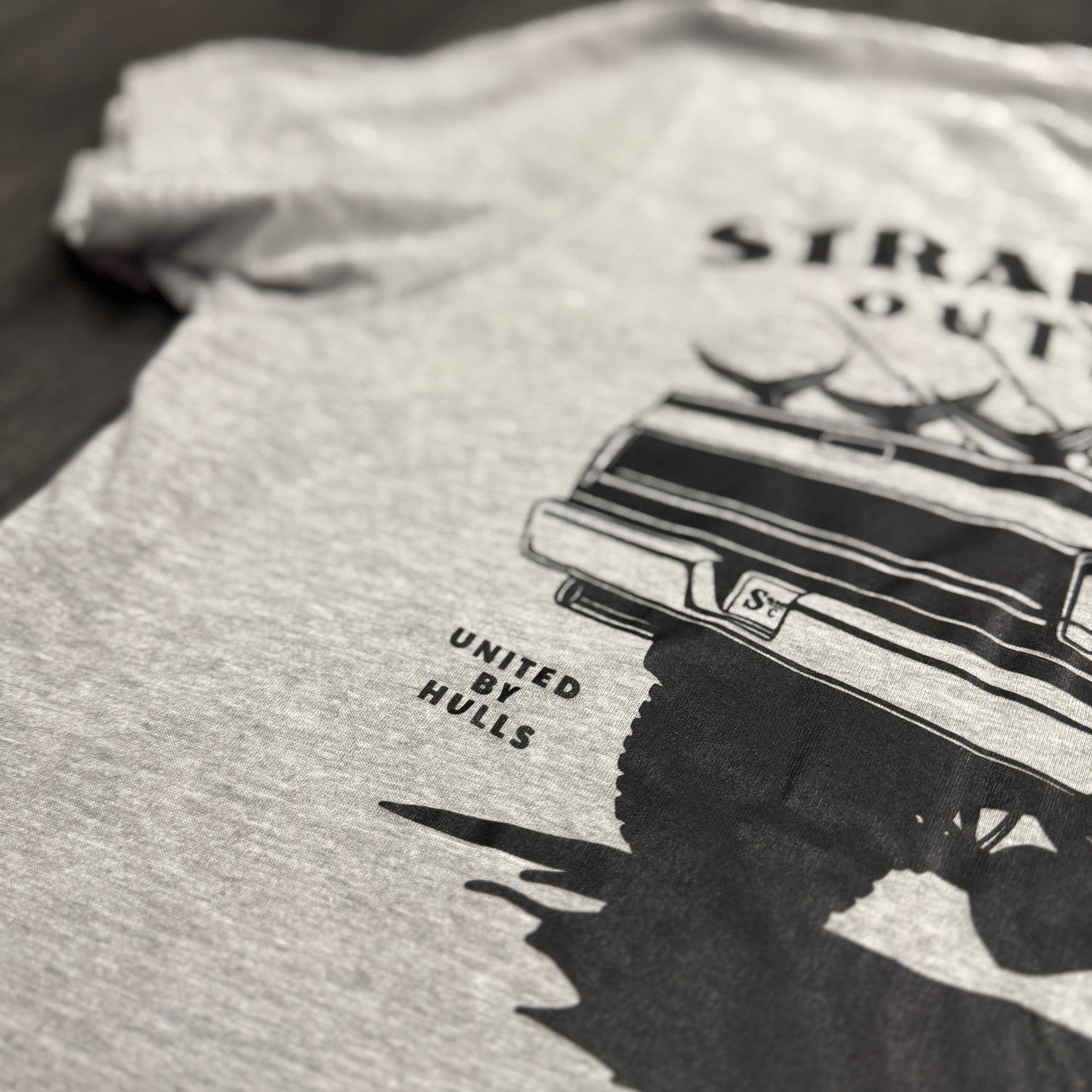 Gray t-shirt with black graphic design of a truck and text on a wooden surface